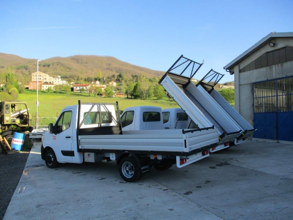 Due camion bianchi con cassone ribaltabile rialzato parcheggiati su un piazzale di cemento di fronte a un edificio, con le montagne sullo sfondo.