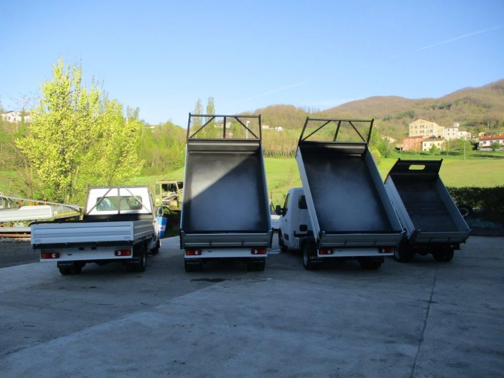 Quattro autocarri ribaltabili parcheggiati in fila su un piazzale di cemento, con tre cassoni sollevati, sullo sfondo di colline e alberi.