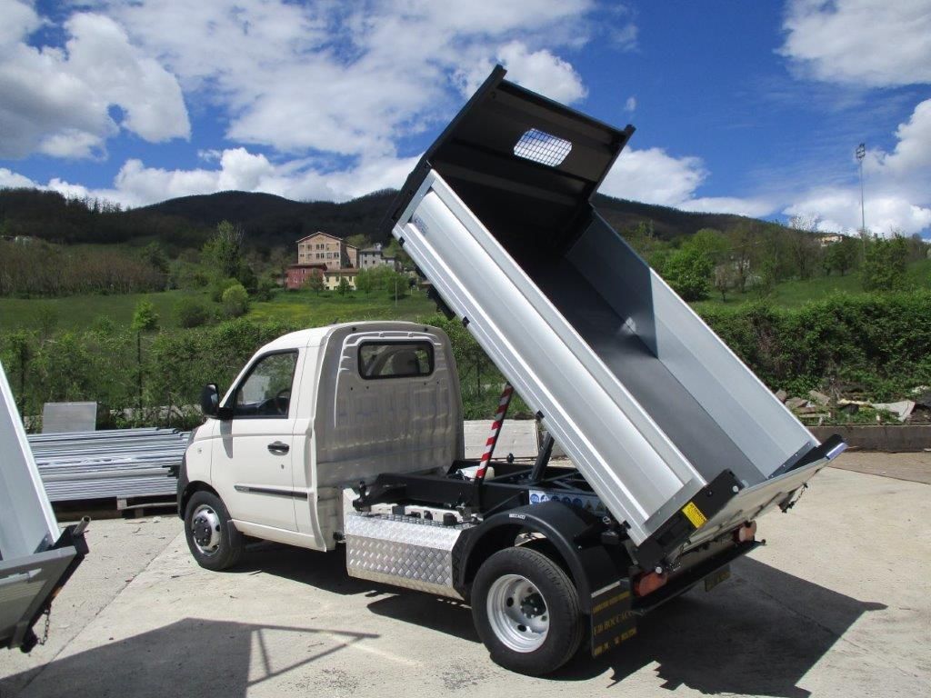 Un camion ribaltabile bianco con il cassone completamente sollevato, parcheggiato all'aperto su un piazzale asfaltato, sullo sfondo di un paesaggio montano.