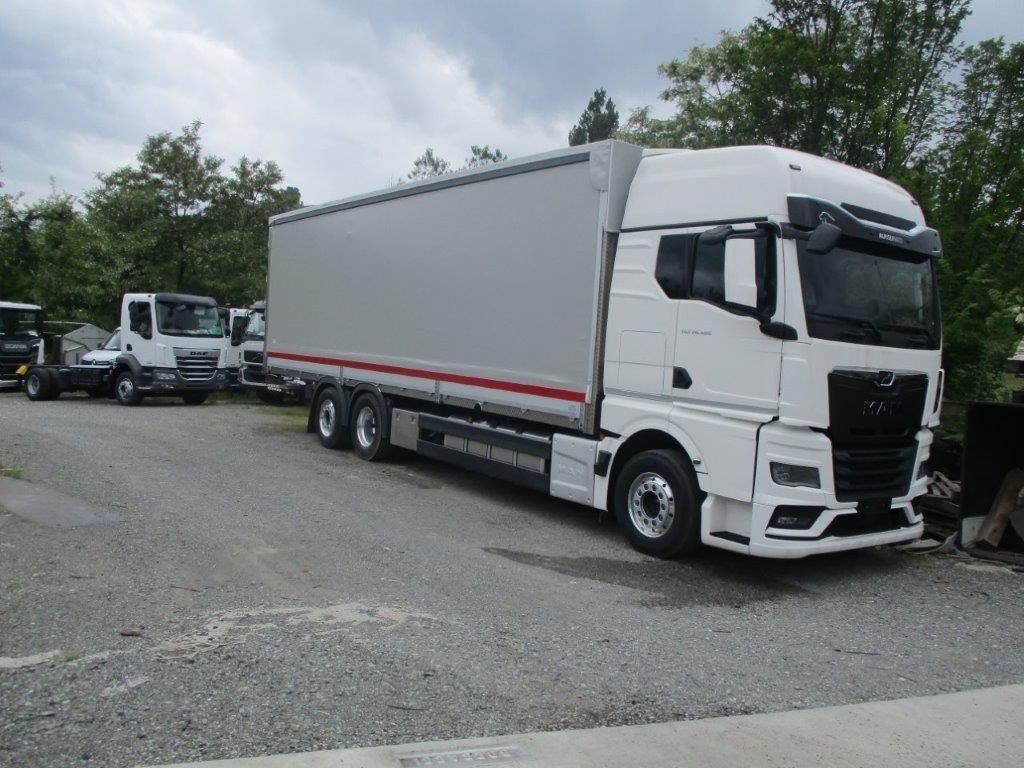 Un camion MAN bianco con rimorchio grigio parcheggiato su un piazzale ghiaioso vicino ad alberi in una giornata nuvolosa.