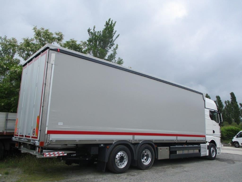 Un camion bianco con un grande rimorchio grigio a sponde piatte parcheggiato all'aperto su un piazzale ghiaioso vicino agli alberi.