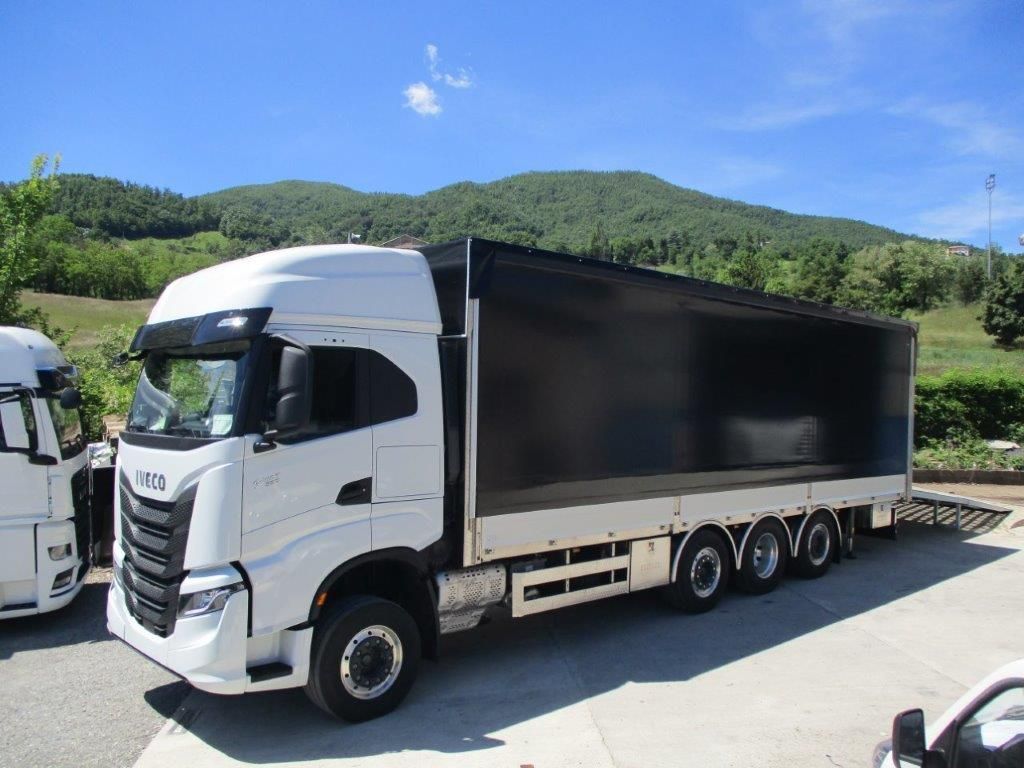 Un camion Iveco bianco con cassone coperto di nero parcheggiato su un piazzale asfaltato, sullo sfondo di verdi colline.
