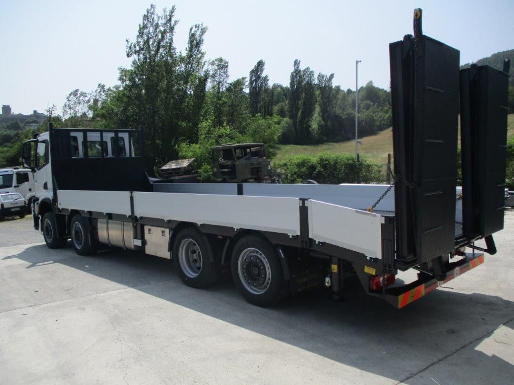 Un camion bianco con pianale e rampe pieghevoli nere sul retro, parcheggiato su un piazzale asfaltato in una giornata di sole.