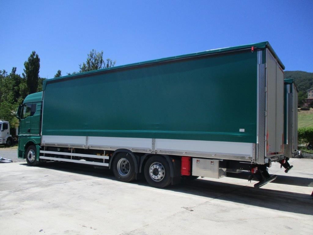 Un semirimorchio telonato verde parcheggiato su un piazzale di cemento sotto un cielo azzurro e limpido.