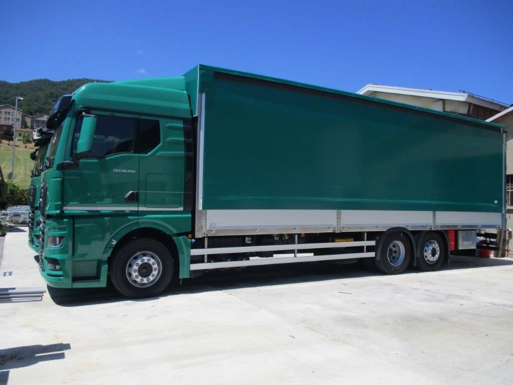 Un camioncino verde per le consegne, adibito a servizio commerciale, è parcheggiato su una superficie di cemento, stagliandosi contro un cielo azzurro e limpido.