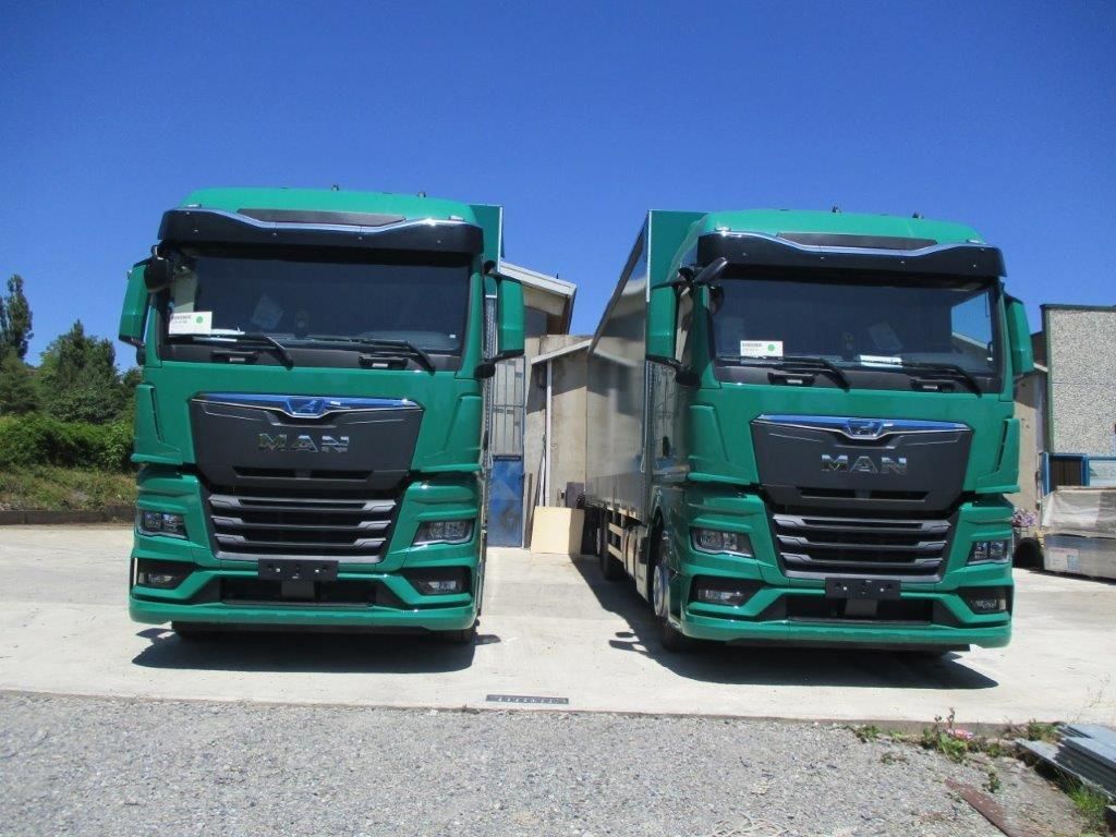 Due camion MAN identici, di colore verde, parcheggiati fianco a fianco in un piazzale ghiaioso sotto un cielo azzurro e limpido.