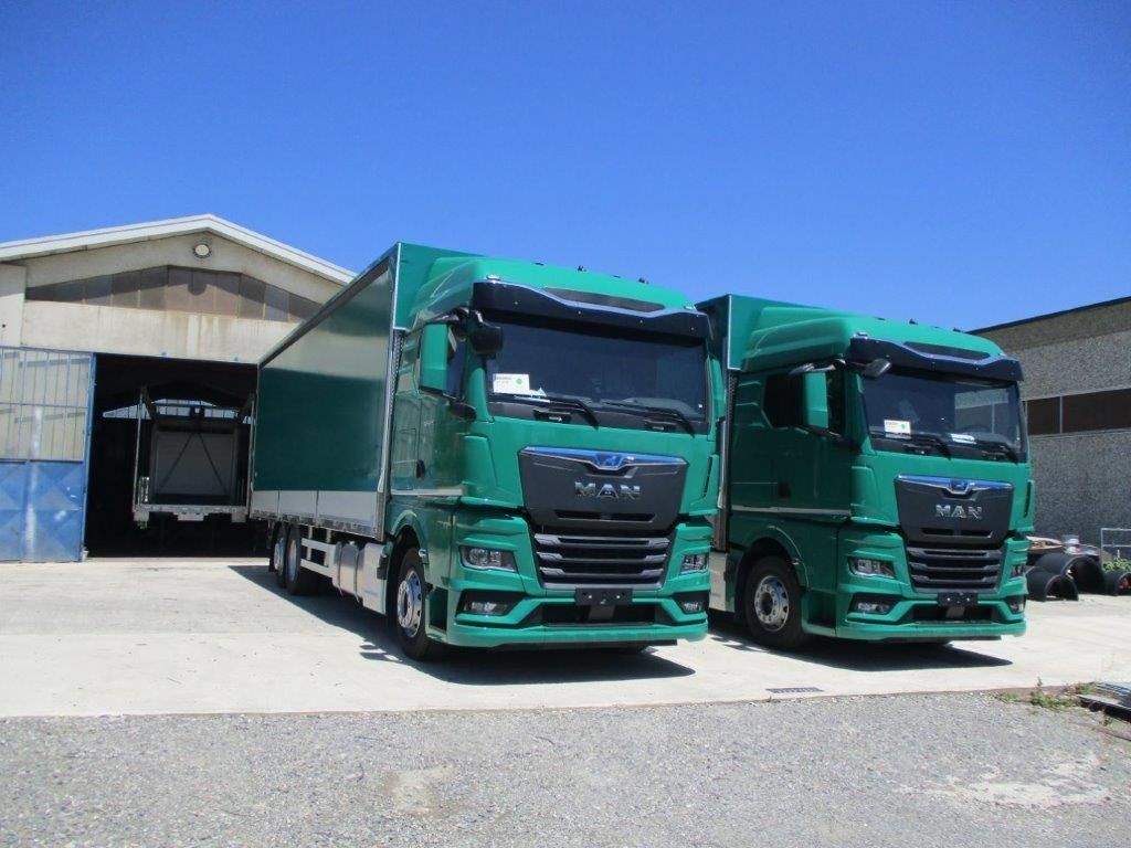 Due autoarticolati MAN verdi parcheggiati fianco a fianco davanti a un magazzino industriale, sotto un cielo azzurro e limpido.