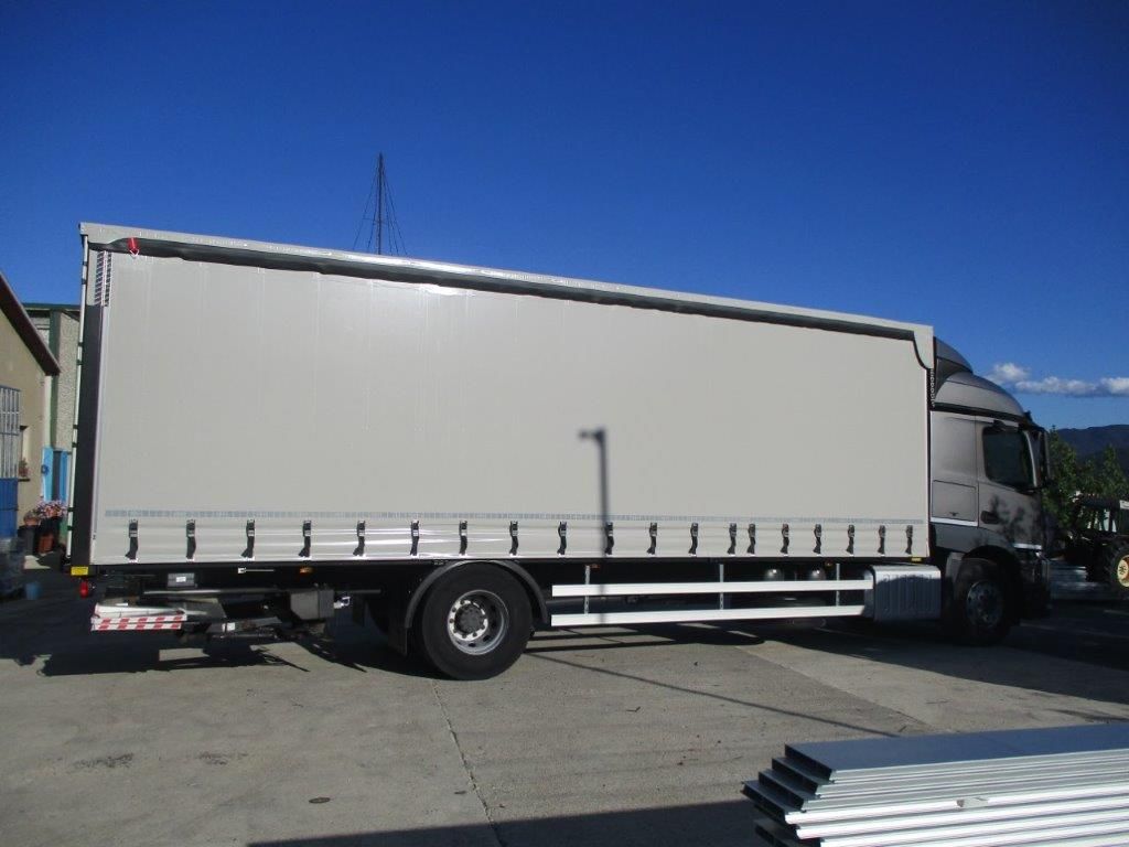 Vista laterale di un furgone commerciale bianco con cassone telonato, parcheggiato su un piazzale asfaltato sotto un cielo azzurro.