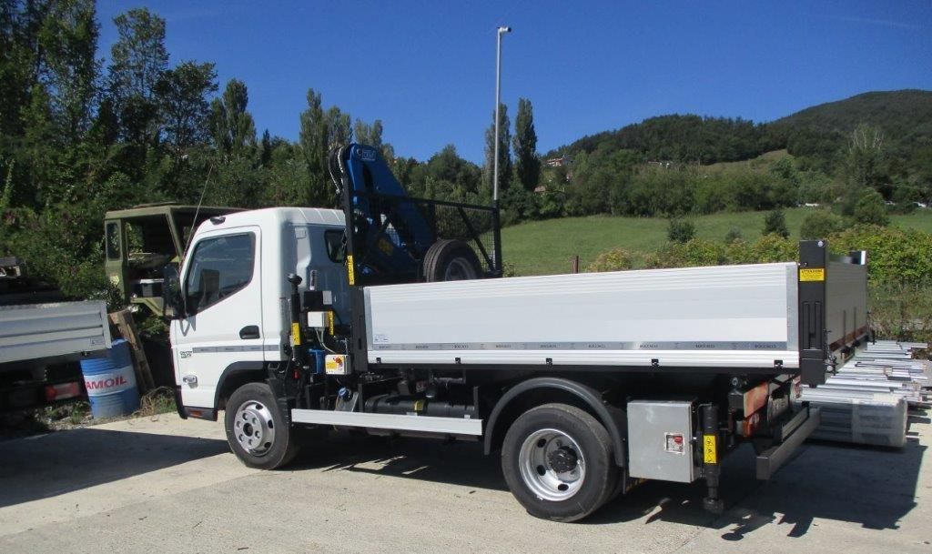 Un camioncino bianco con gru montata, parcheggiato in un piazzale ghiaioso all'aperto in una giornata di sole.
