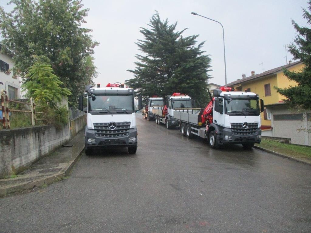 Tre camion Mercedes-Benz bianchi con pianale e gru rosse parcheggiati su una strada asfaltata tra edifici e alberi.