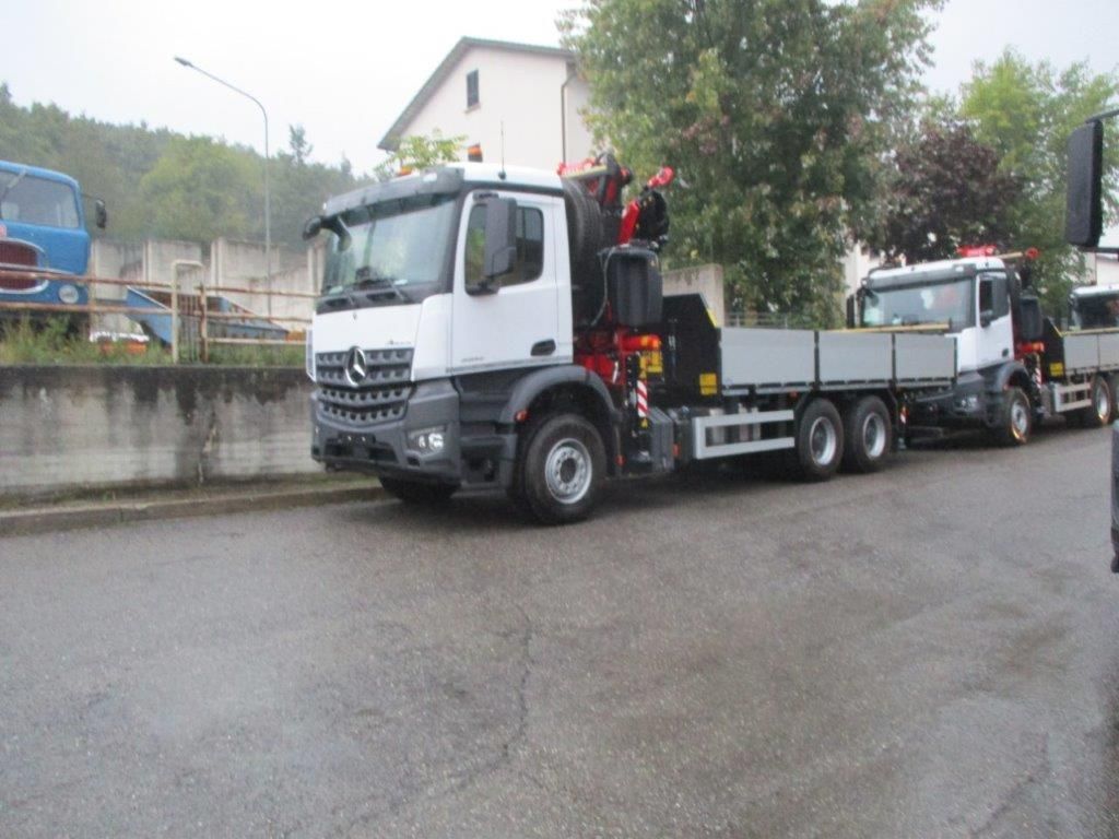 Due camion Mercedes-Benz bianchi con pianale e gru, parcheggiati su un piazzale asfaltato vicino a un edificio.