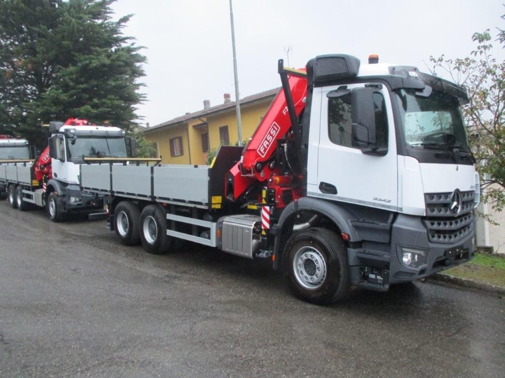 Due camion Mercedes-Benz bianchi con pianale e gru di carico rosse parcheggiati su una strada asfaltata vicino a un edificio.