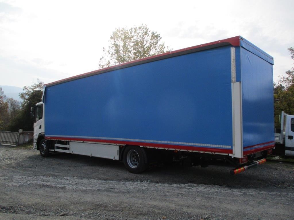 Un grosso camion blu con rimorchio è parcheggiato su un piazzale ghiaioso vicino ad alberi, sotto un cielo nuvoloso.