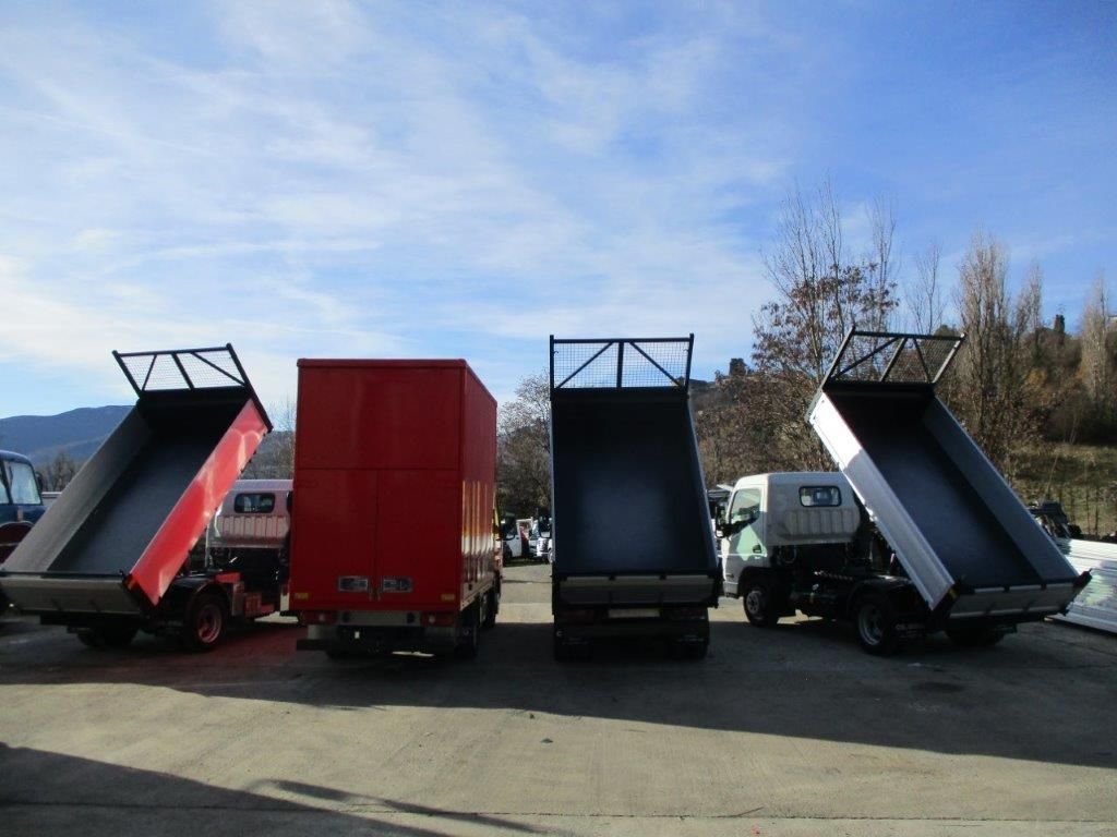 Quattro camion parcheggiati all'esterno, con tre cassoni ribaltabili sollevati, in un piazzale all'aperto sotto un cielo azzurro e soleggiato.