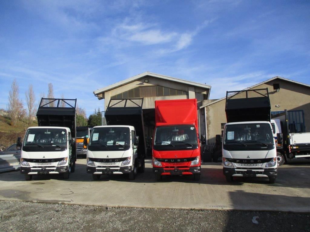 Quattro autocarri ribaltabili, tre bianchi e uno rosso, parcheggiati in fila davanti a un edificio sotto un cielo azzurro.