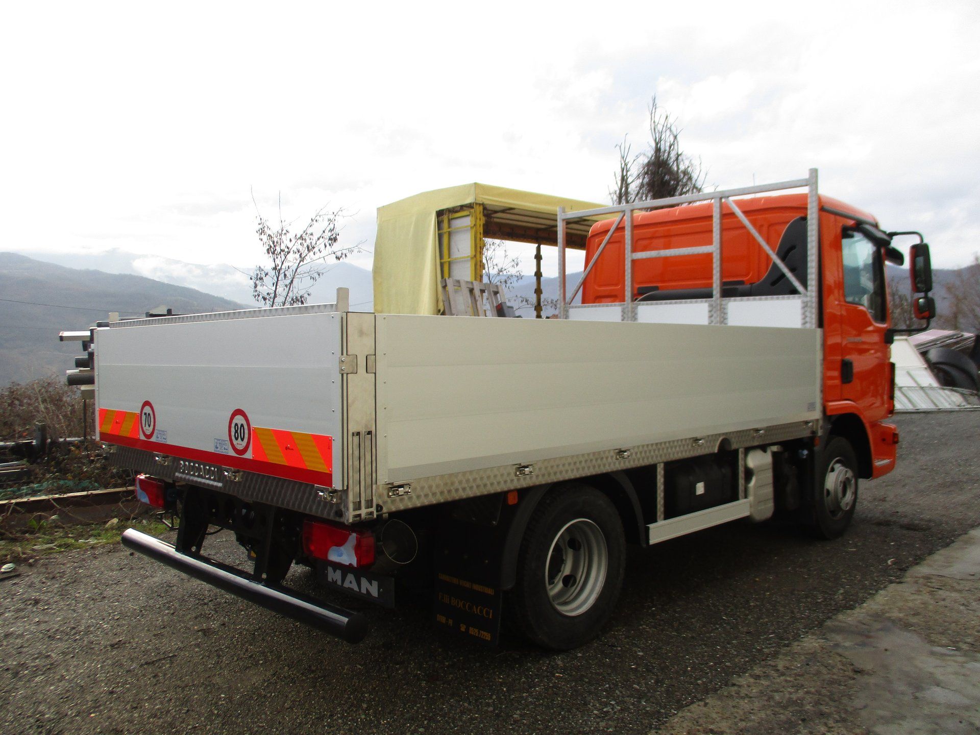 Camion MAN arancione con pianale di carico bianco, parcheggiato su una superficie ghiaiosa all'aperto con alberi e colline sullo sfondo.