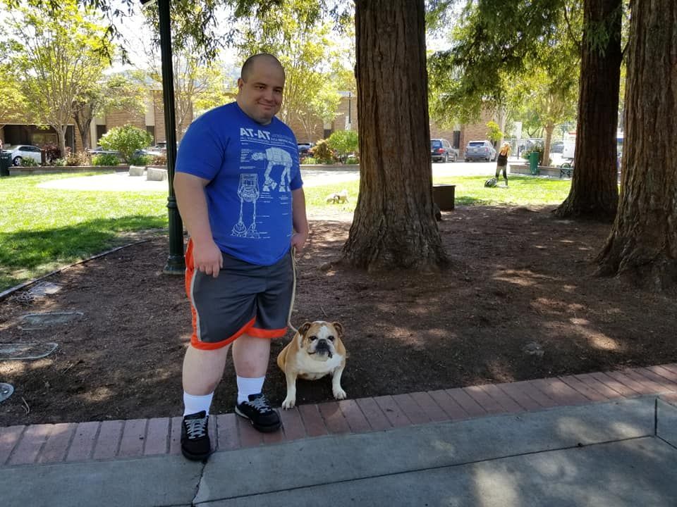 Man in blue shirt and bulldog dog standing by trees outdoors.