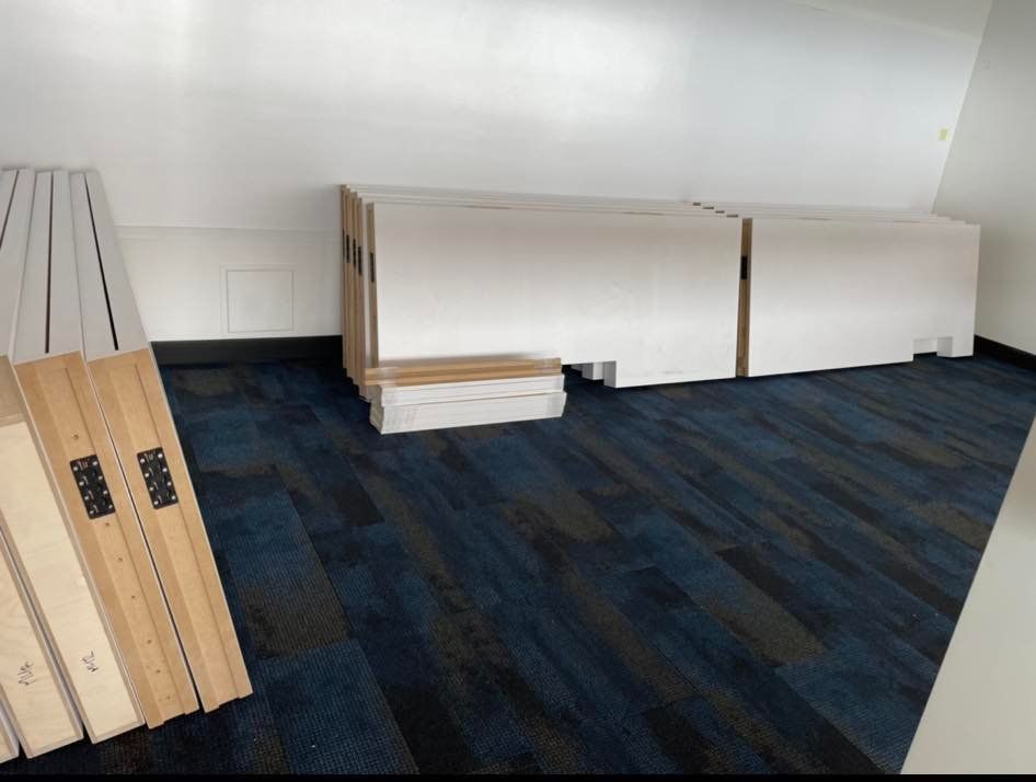 Des meubles blancs reposent sur un tapis bleu dans une pièce. Des panneaux de bois sont appuyés contre un mur.