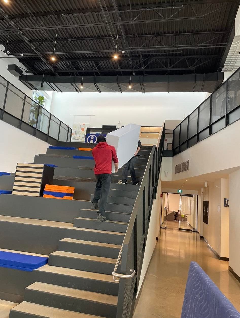 Deux personnes transportant une grande boîte blanche dans des escaliers à plusieurs niveaux dans un immeuble moderne.