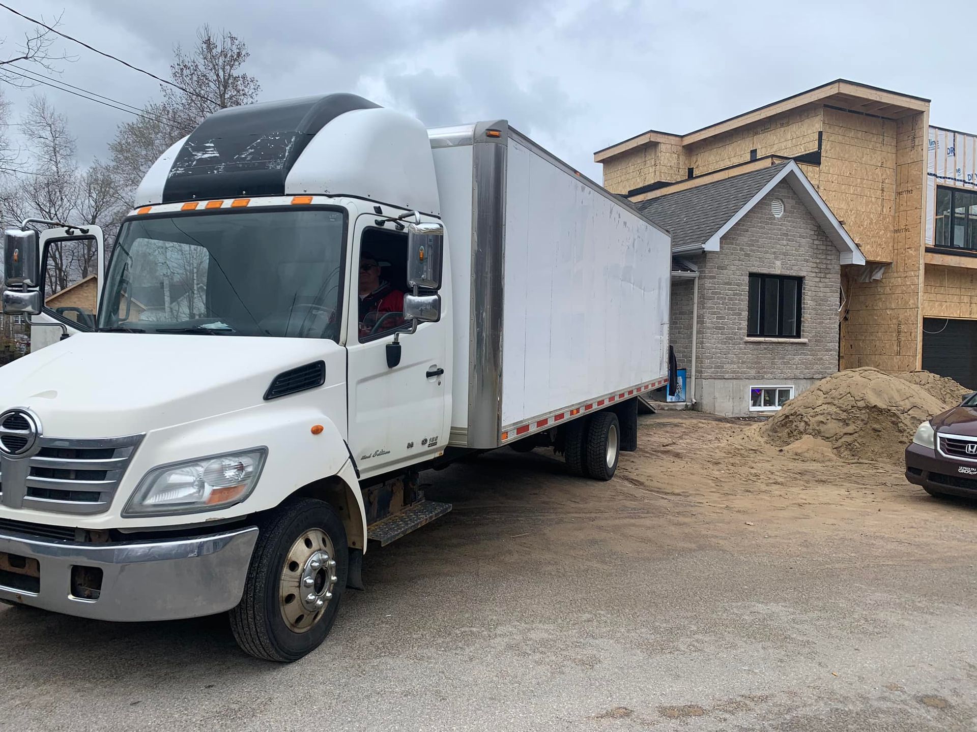Camion blanc garé à côté d'une maison en construction ; tas de sable brun ; ciel gris.