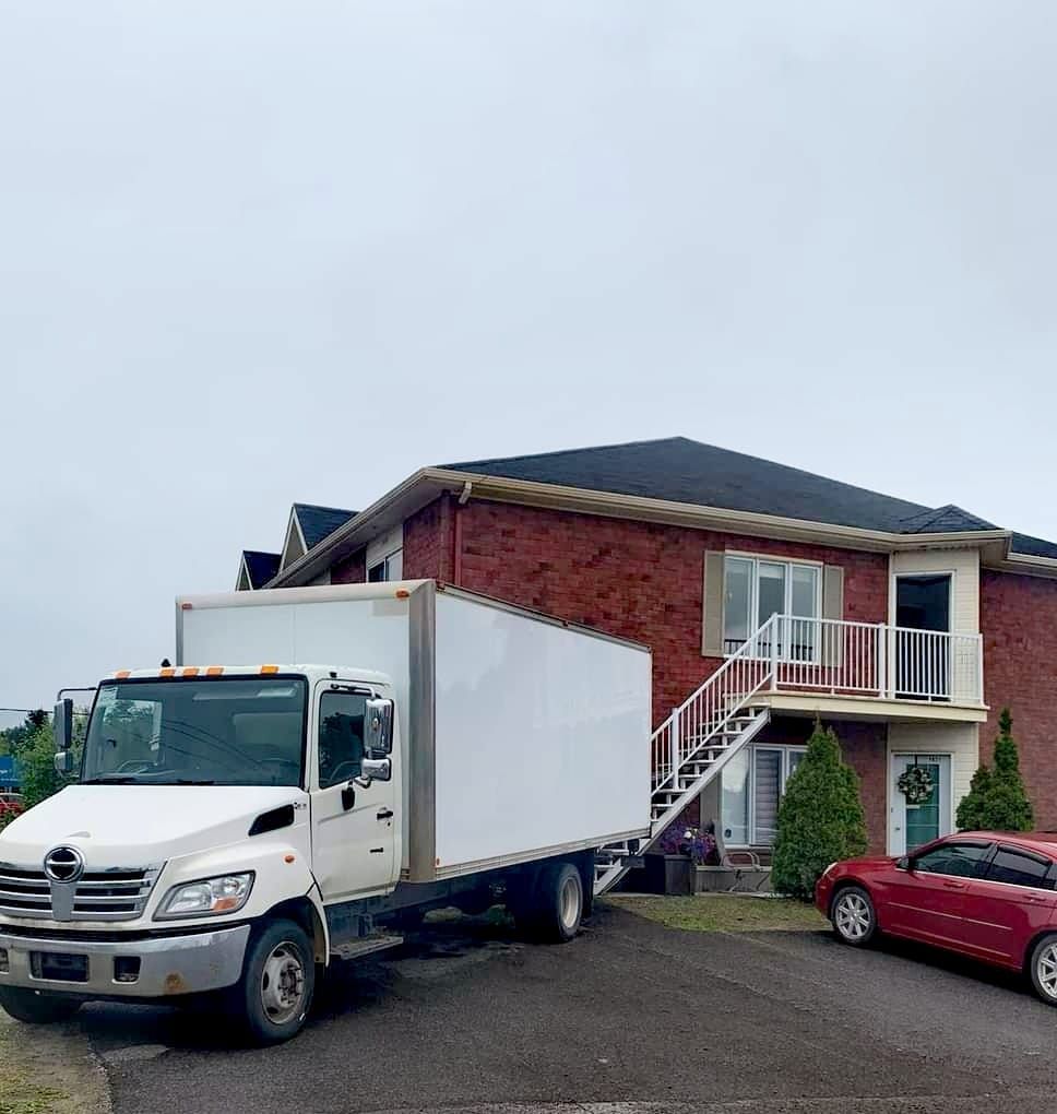 Un camion de déménagement blanc garé devant une maison en briques à deux étages ; une voiture rouge garée dans l'allée.