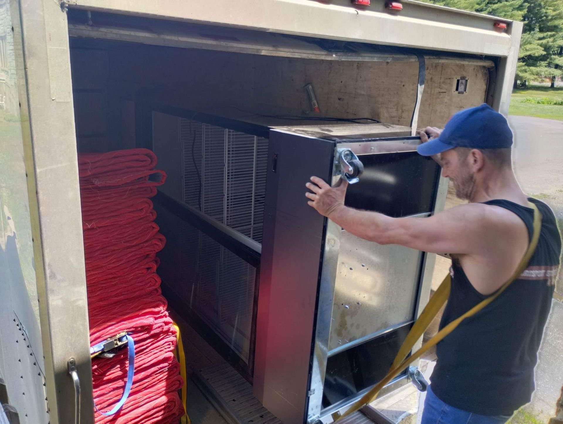 Un homme charge un gros appareil sombre dans un camion ; sangles rouges à gauche.