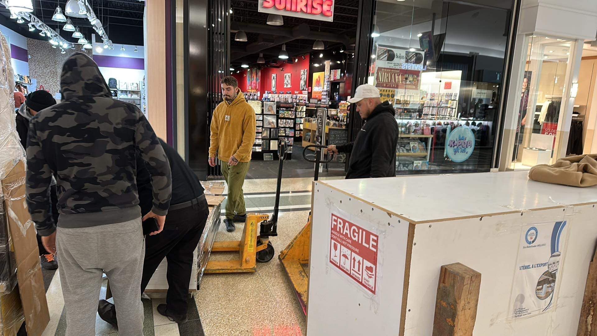 Des gens déplacent une grande caisse à l'extérieur d'un magasin appelé Sunrise dans un centre commercial.