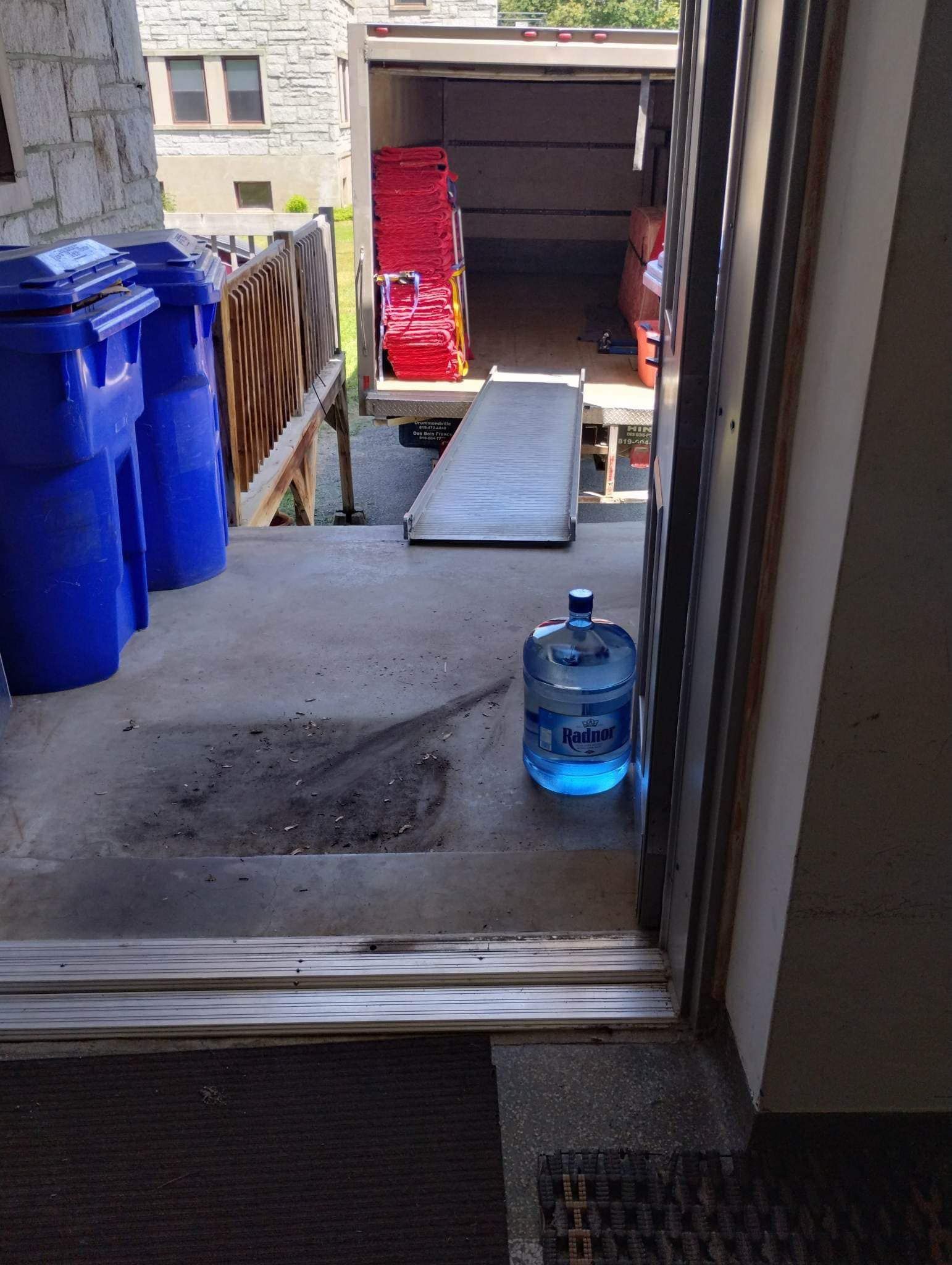 Une personne charge un camion avec des cartons sur une rampe ; des poubelles bleues à gauche, une cruche d'eau au milieu.