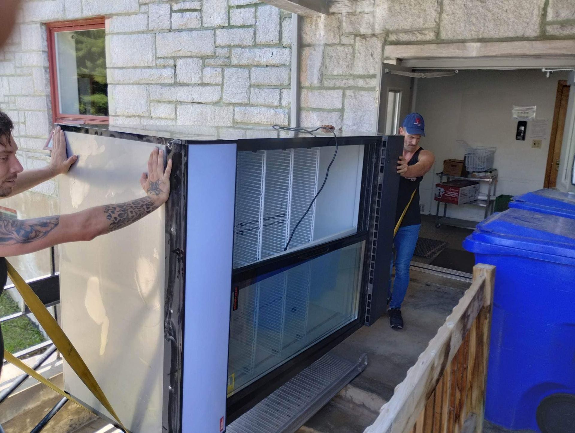 Deux personnes déplacent une grande glacière à façade vitrée hors d'un garage.