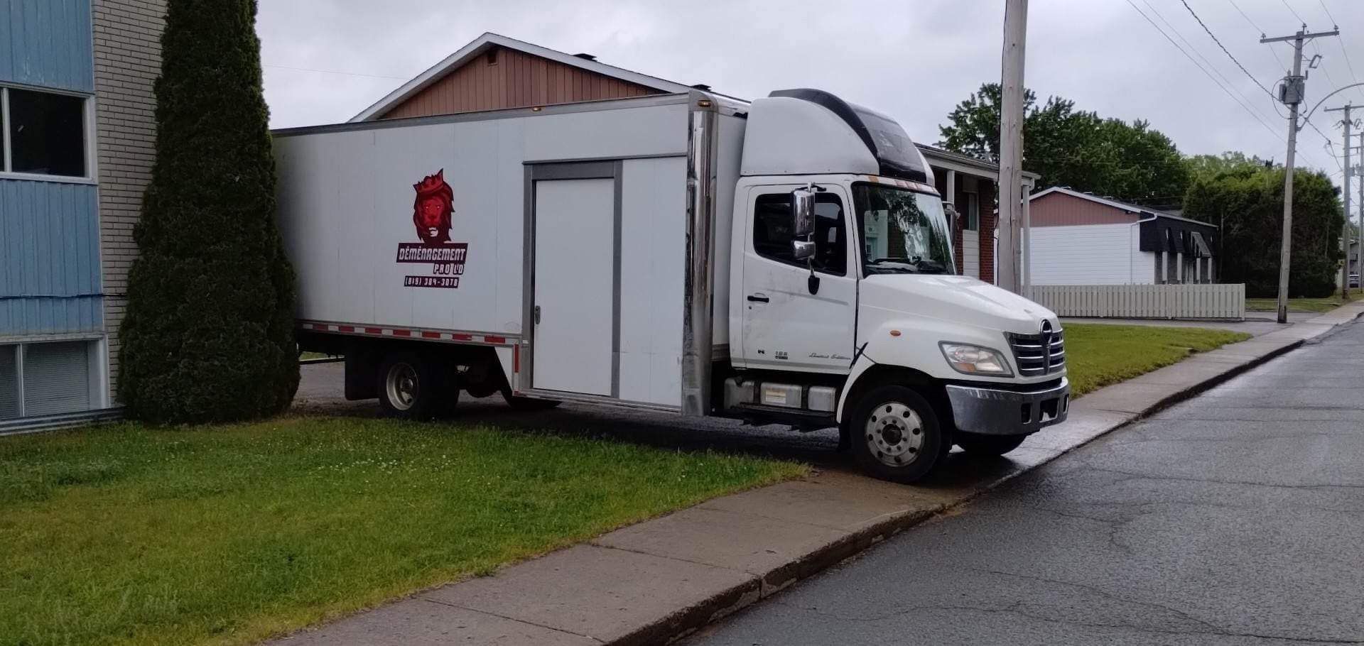 Un camion-caisse blanc sur le trottoir, à côté d'un bâtiment. Le camion porte un logo sur le côté.