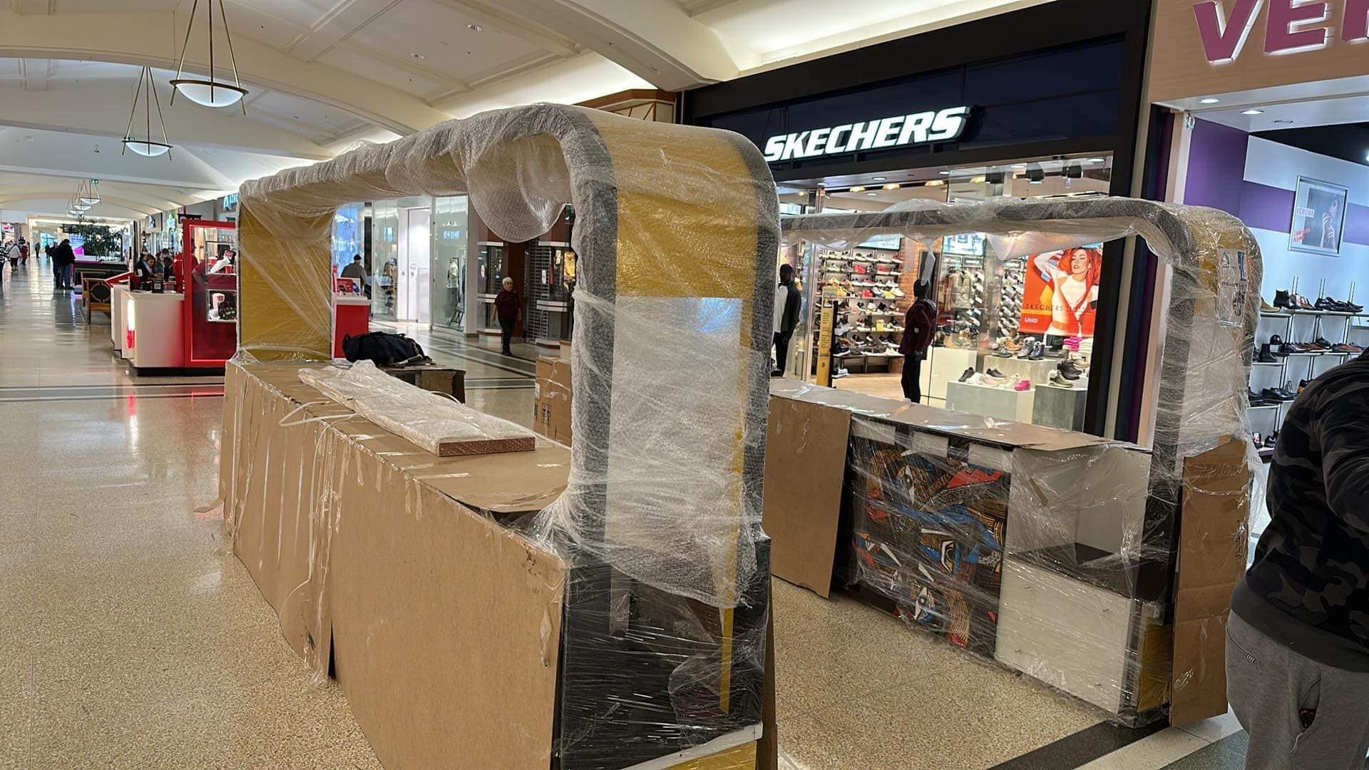 Kiosque de centre commercial enveloppé de plastique et de carton, avec des magasins et des acheteurs visibles en arrière-plan.