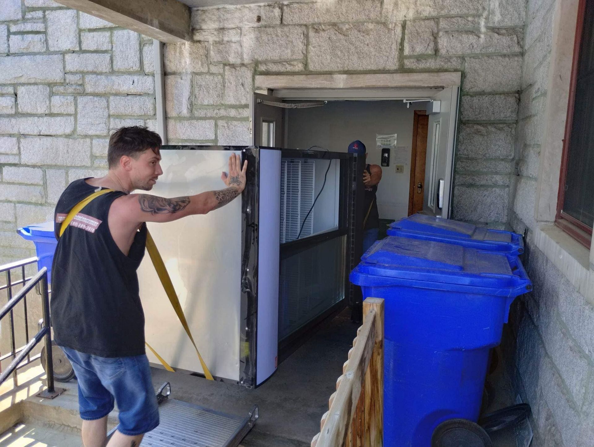 Deux déménageurs transportent un objet volumineux, probablement un matelas, par une porte. L'un pousse, l'autre guide.