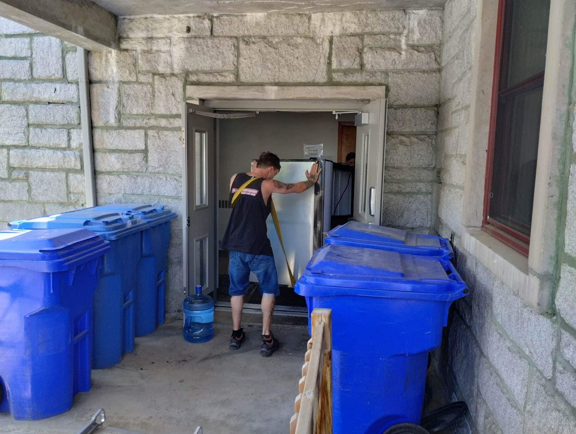 Un homme s'efforce de déplacer un gros appareil à travers une porte, entouré de bacs de recyclage bleus et de murs en pierre.