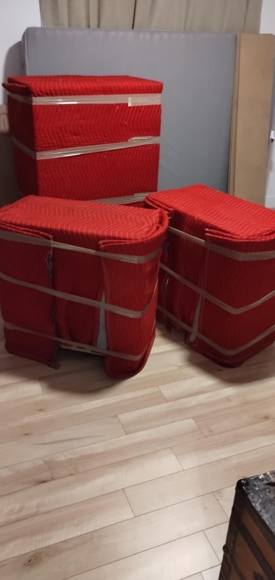 Trois objets rouges, emballés et scotchés, peut-être des meubles, sur un plancher en bois, devant une surface grise.