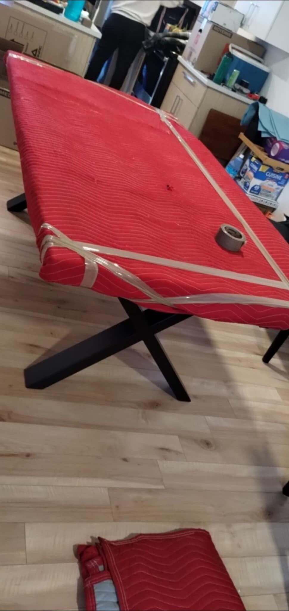 Une table rouge enveloppée dans une housse de protection et scotchée pour le transport. Un rouleau de ruban adhésif est posé dessus.