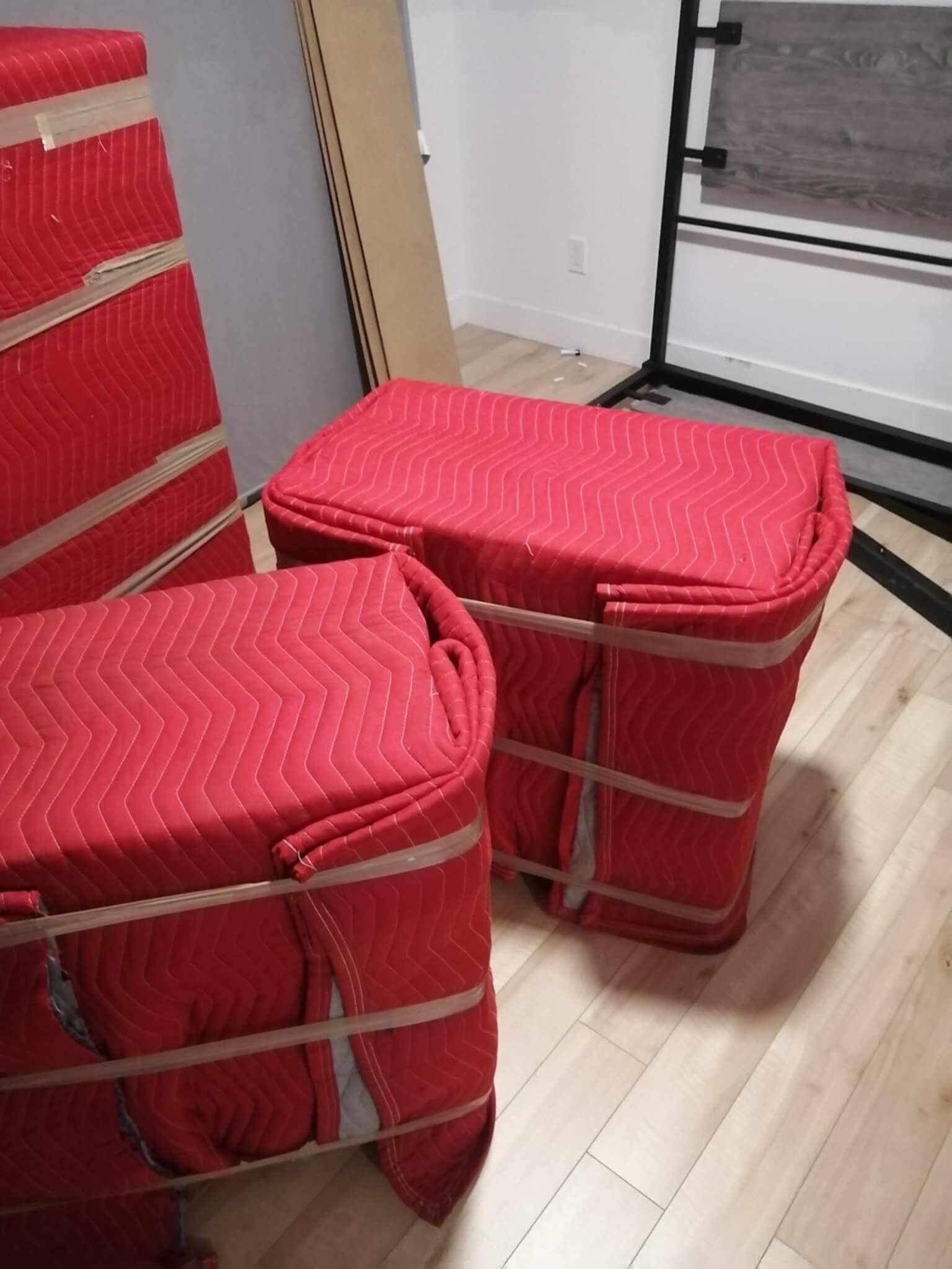 Deux objets rembourrés rouges enveloppés dans des couvertures de déménagement, fixés avec des sangles, sur un plancher en bois.