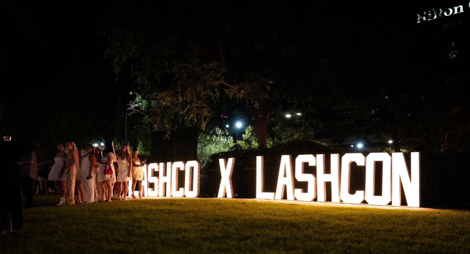 A group of people are standing in front of a large sign that says lashcon.
