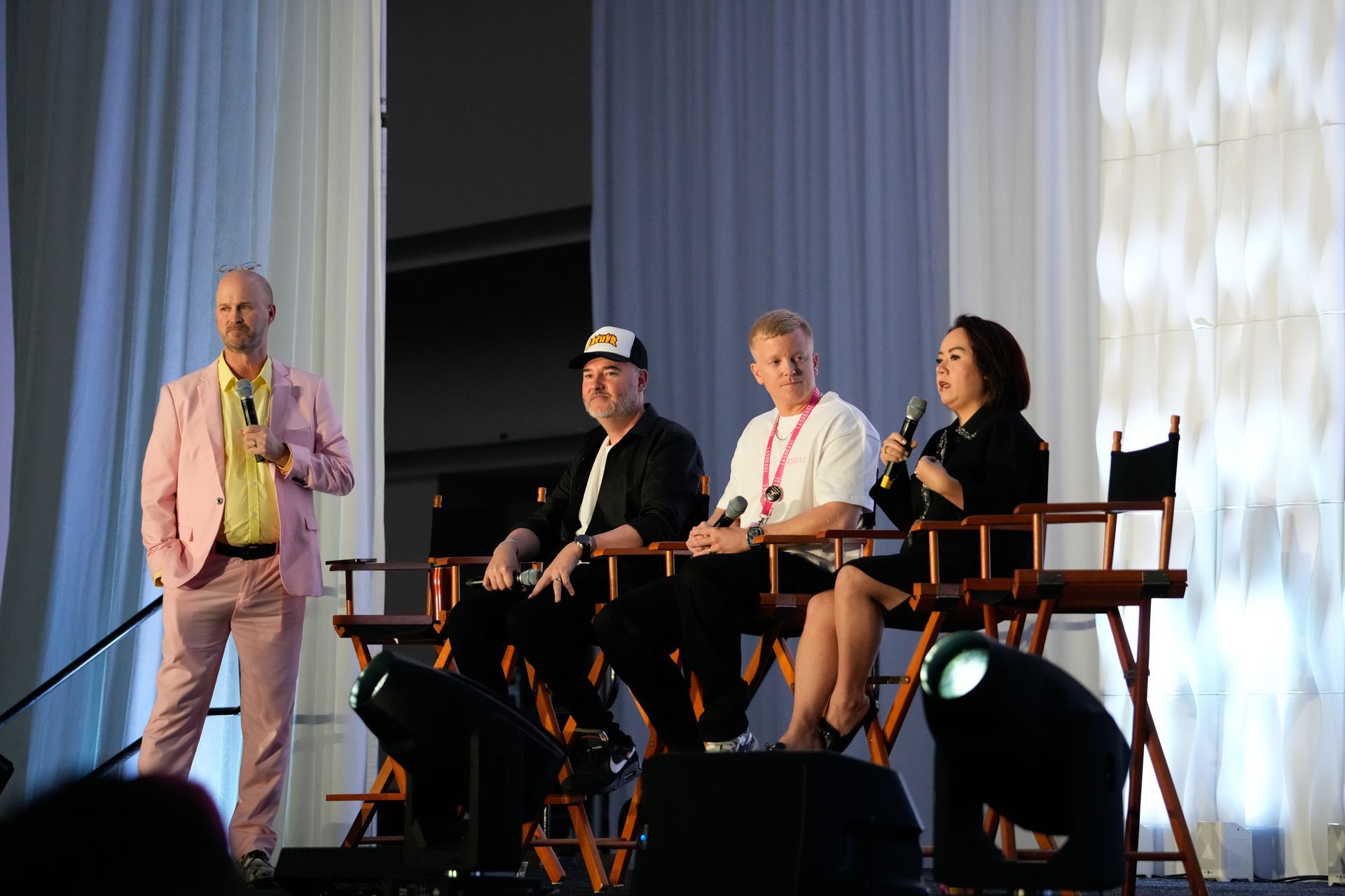 A group of people sitting in chairs on a stage with a man in a pink suit speaking into a microphone.