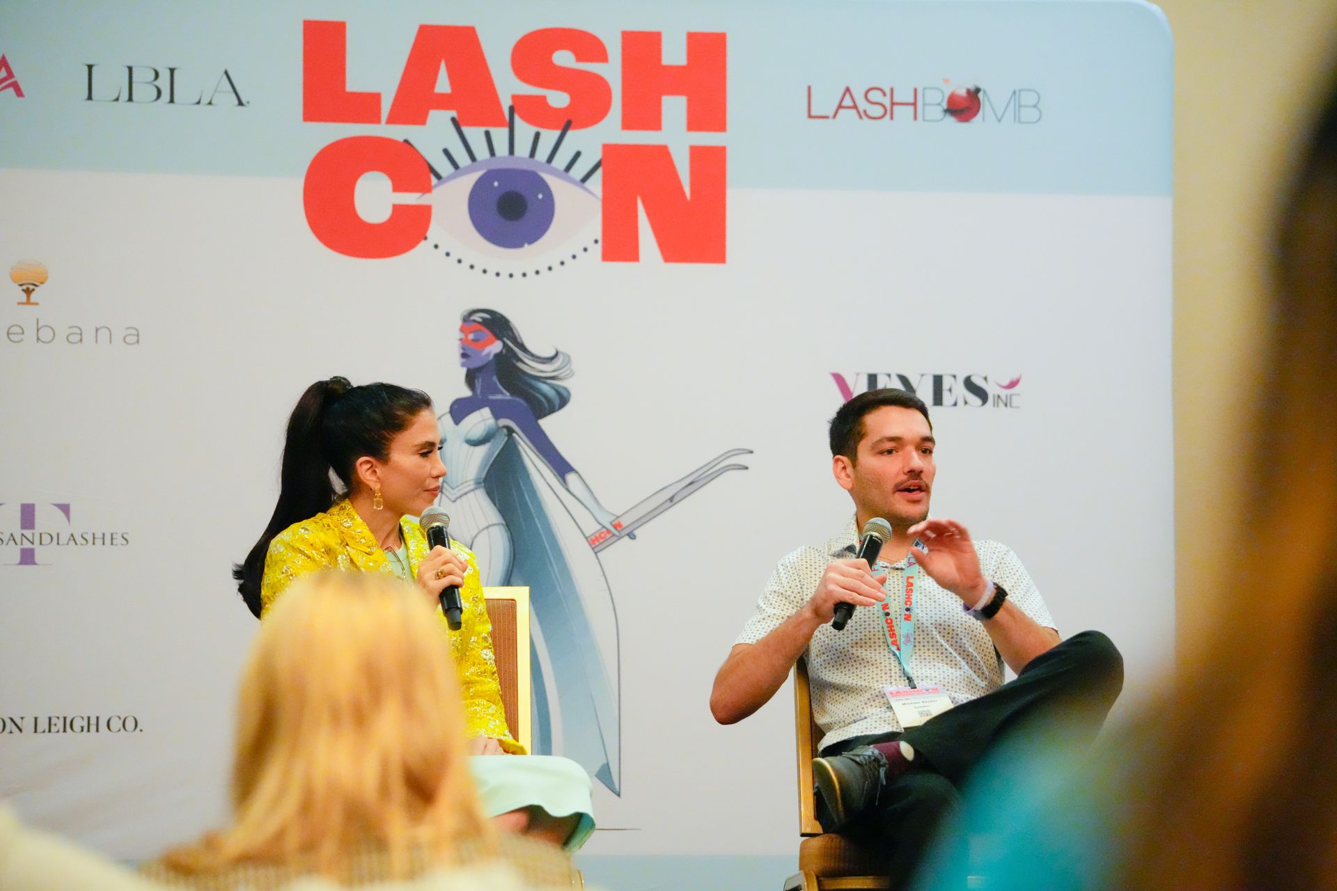 A man and a woman are sitting in front of a lash con banner.