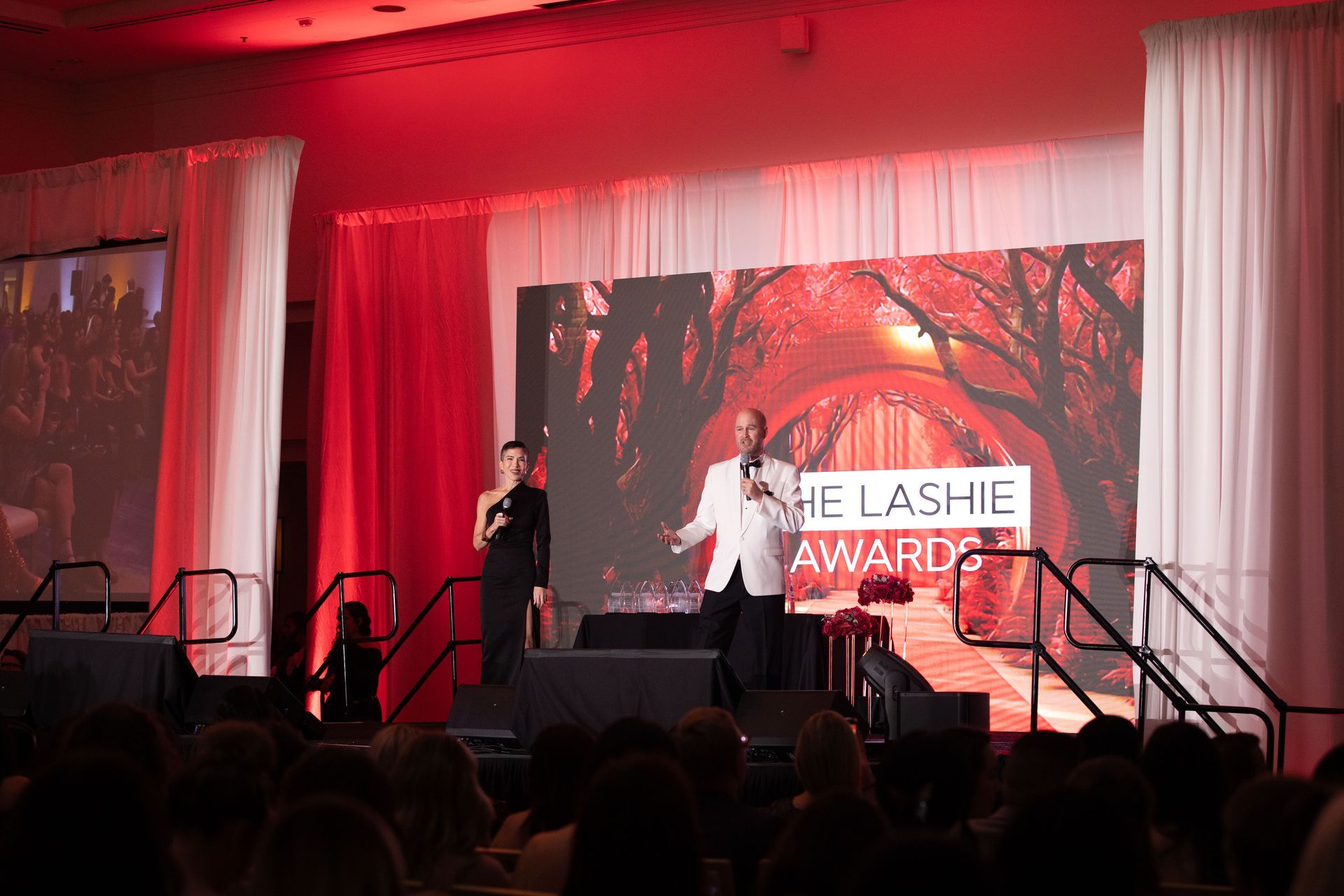 Two presenters stand on a stage in front of a screen reading 