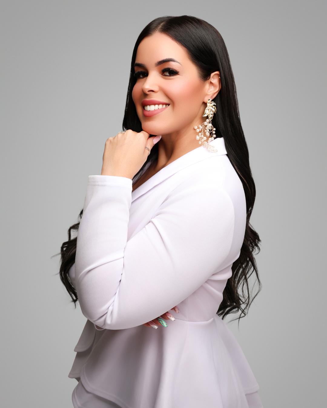 A woman wearing a white jacket and earrings is standing with her hand on her chin.