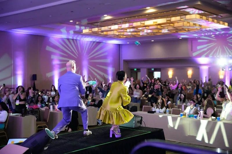 Two people on stage in front of a cheering crowd, purple lighting, festive event.