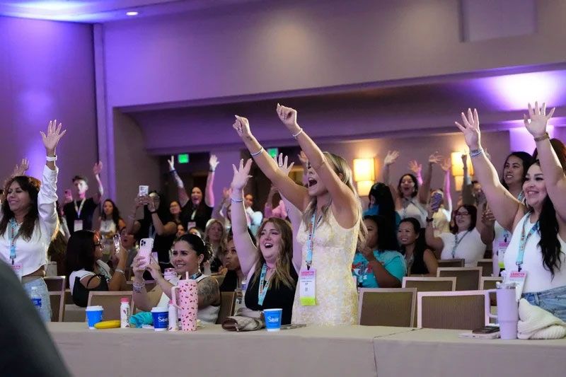 People raising hands in a conference room with a purple hue. Women are in the front row cheering.