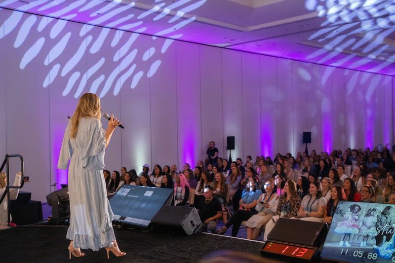 Woman speaking onstage to a large audience at a conference; purple lighting, screens, and speakers.