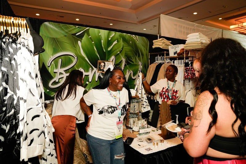 Clothing booth at a fair with people browsing and interacting. Green banner with logo and clothes on display.