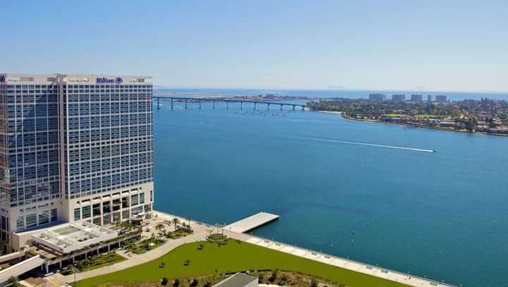 High-rise building next to a body of water with a bridge in the distance, under a blue sky.