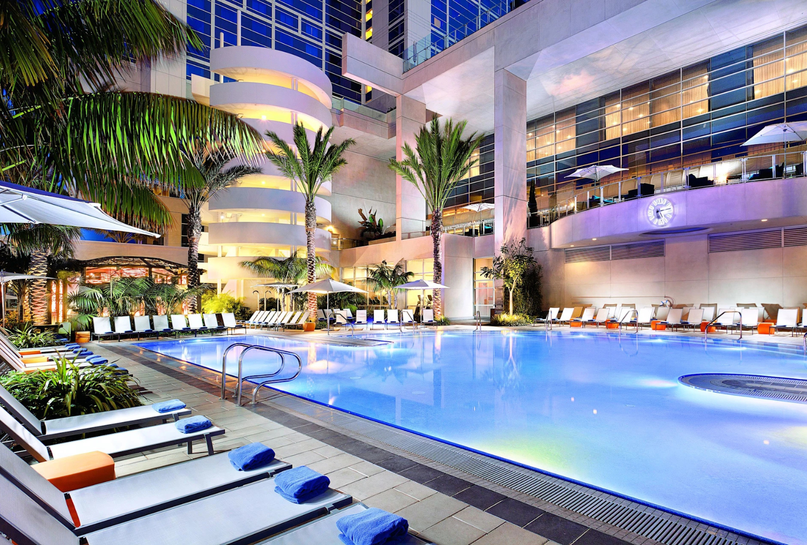 A luxury hotel swimming pool at night, surrounded by lounge chairs, palm trees, and modern architectural lighting.