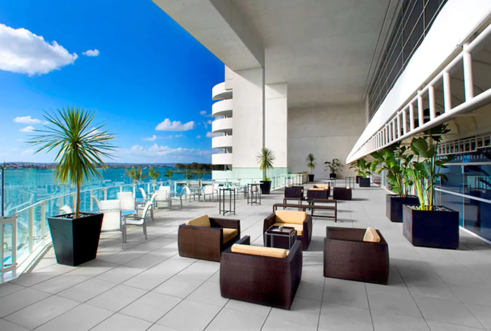 A sunny outdoor terrace with modern dark furniture, potted plants, and a glass railing overlooking the sea.