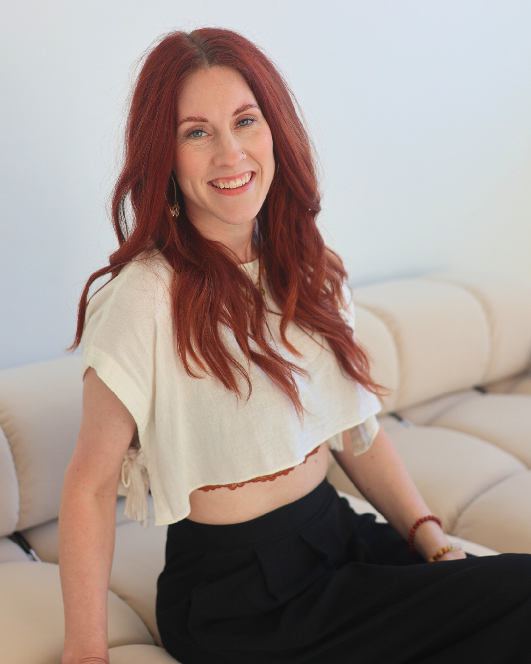 A woman with red hair is sitting on a couch wearing a white crop top and black pants.
