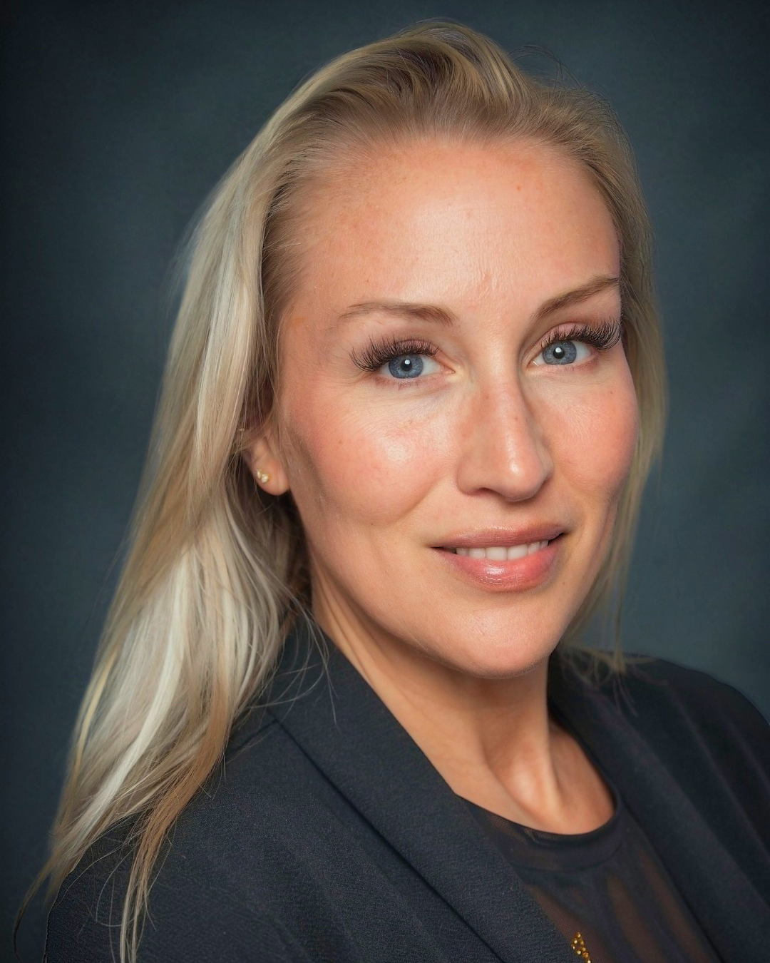 A woman with blonde hair and blue eyes is wearing a black jacket.
